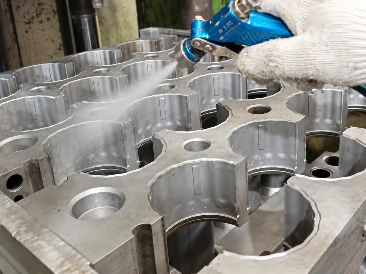 A gloved hand using a blue spray gun over open metal mold cavities